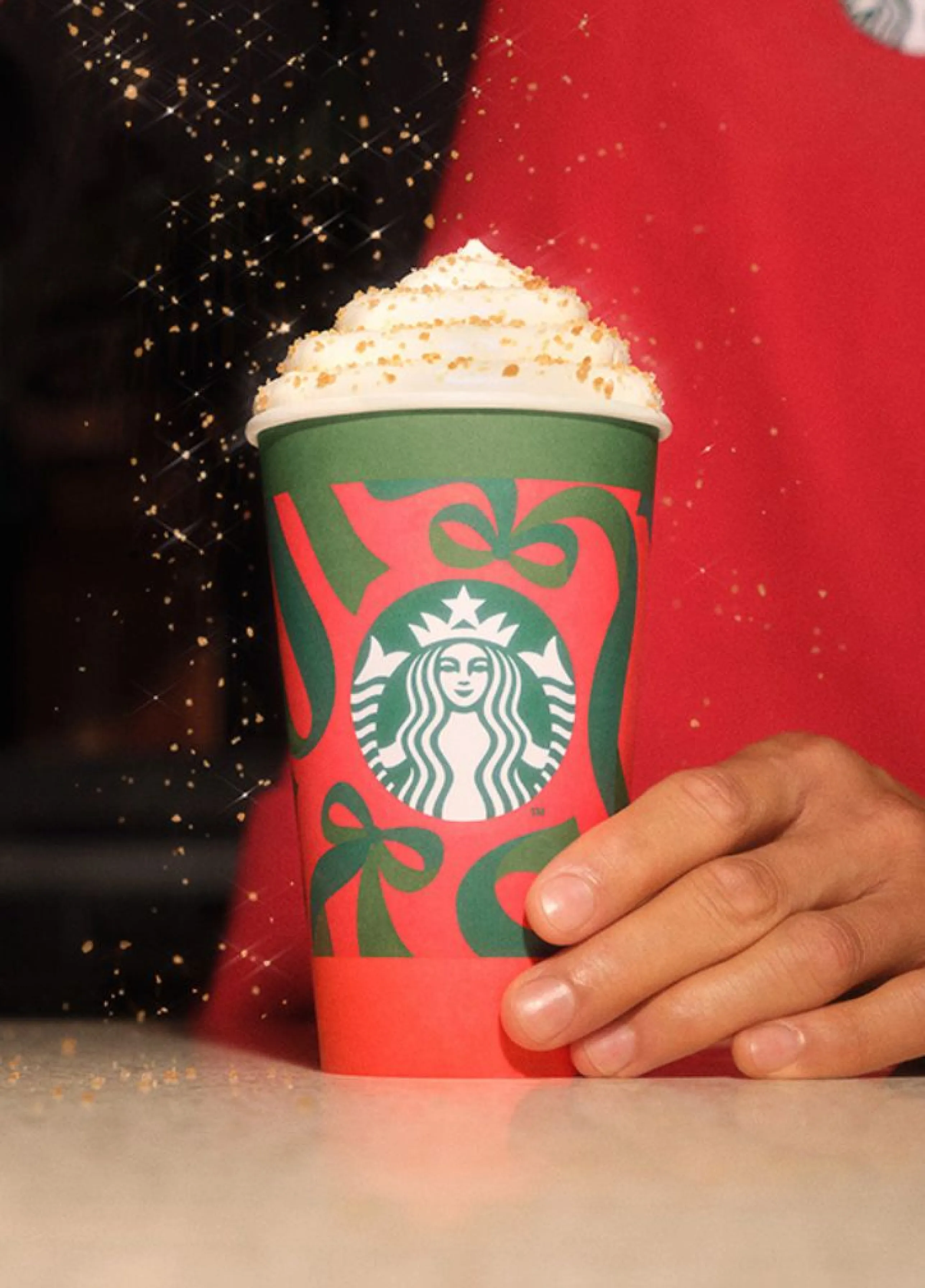Ruka drží červený vianočný pohár Starbucks so vzorom zelených stúh. Pohár je plný nápoja so šľahačkou, na ktorú sa sype cukrová posýpka