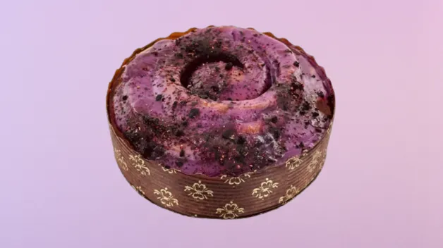 Kulatý pečený dezert s výraznou fialovou polevou a tmavými drobky na povrchu, uložený v hnědém papírovém košíčku se zlatým květinovým vzorem, na jemném růžovo‑fialovém pozadí.