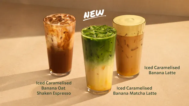 obrázok predstavuje tri nové ľadové nápoje Starbucks z kolekcie Caramelised Banana na teplom béžovom pozadí. Zľava sú to: tmavý espresso nápoj s ovsených mliekom, živozelené matcha latte s banánovou vrstvou a zlatisto-karamelové banánové latte. Nad stredným pohárom je výrazný nápis „NEW".