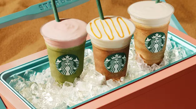 Tri ľadové nápoje Starbucks stoja v nádobe naplnenej ľadom položenej na piesku. Farebné nápoje s penou a slamkami navodzujú letnú plážovú atmosféru.