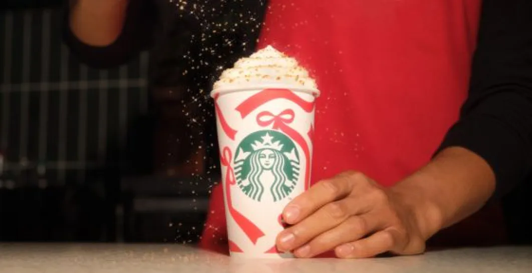 A barista sprinkles gold sprinkles on whipped cream in a red and white Starbucks Christmas cup